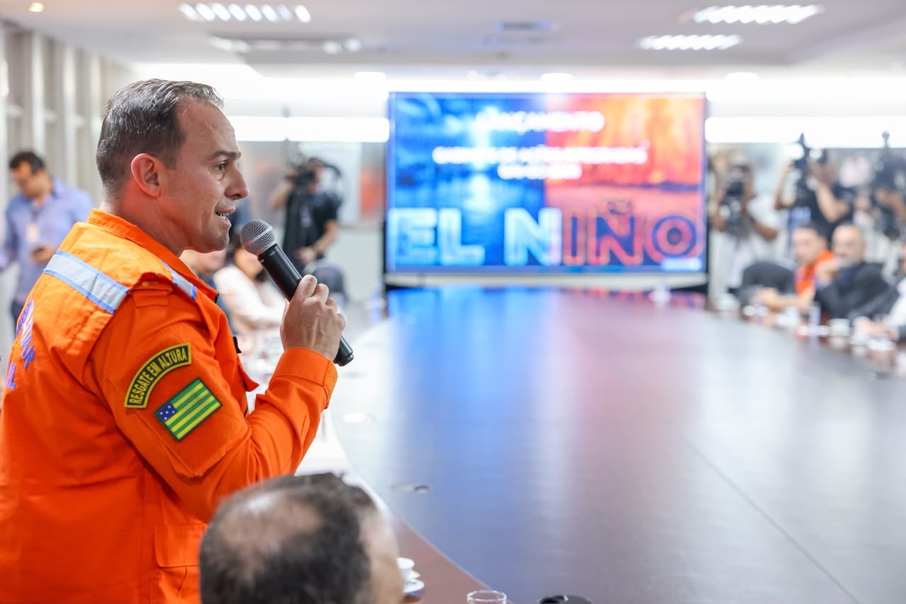 Imagem ilustrativa da reunião com autoridades, bombeiros e defesa civil, e outras entidades estaduais sobre o combate aos impactos do El Niño e queimadas. Em foco na imagem, um bombeiro fala ao microfone.